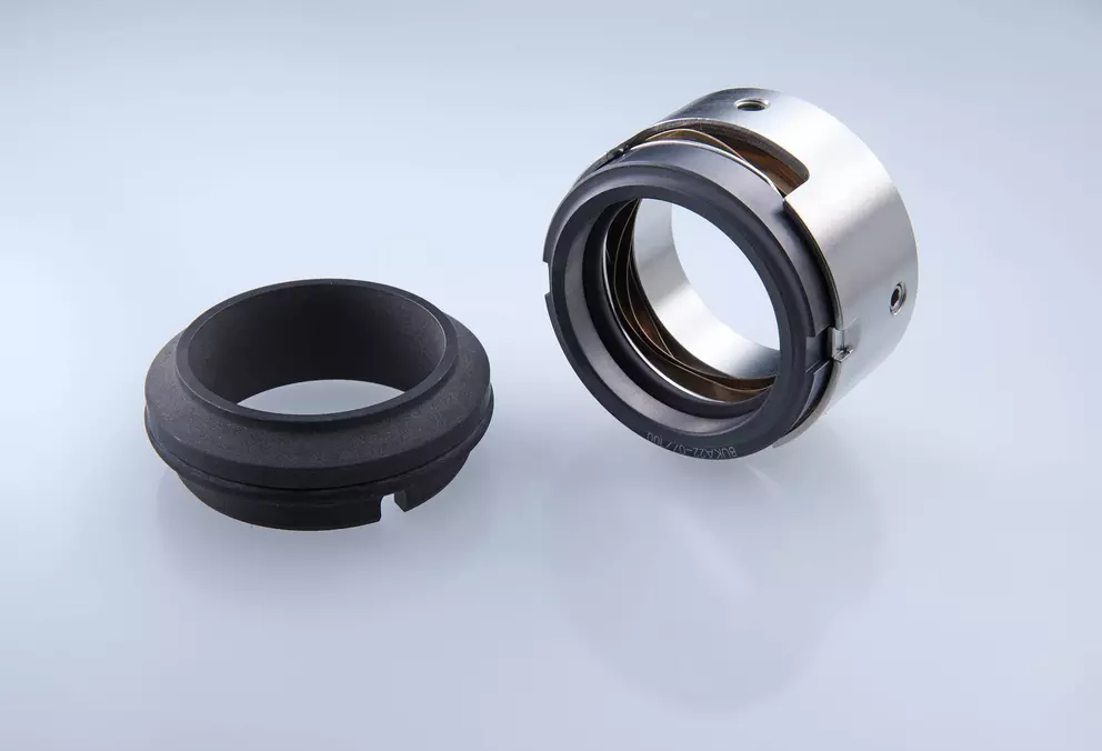 A clean studio photograph of two disassembled industrial mechanical seal components: a black, ribbed elastomer element and a shiny, metallic ring assembly with a black rubber edge and internal springs.,
