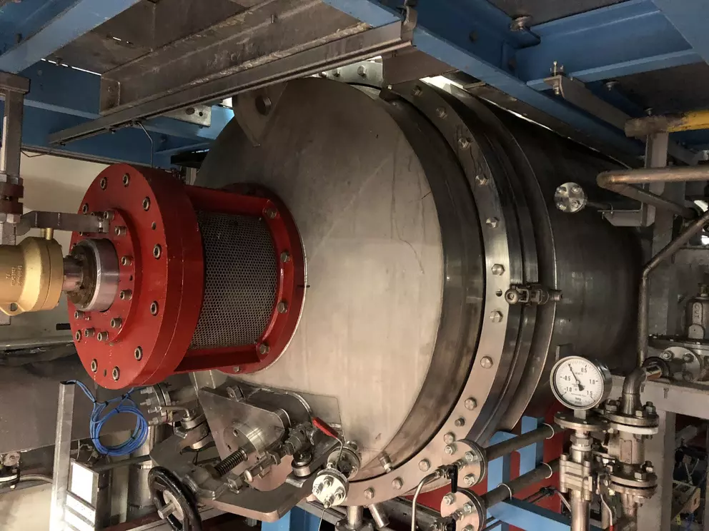 Industrial equipment featuring a large, stainless steel cylindrical vessel with a prominent red flanged attachment and mesh filter, surrounded by piping and gauges.