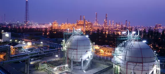 Night-time view of a large petrochemical complex or oil refinery, featuring two massive spherical storage tanks in the foreground and a brightly lit network of towers and stacks in the background.