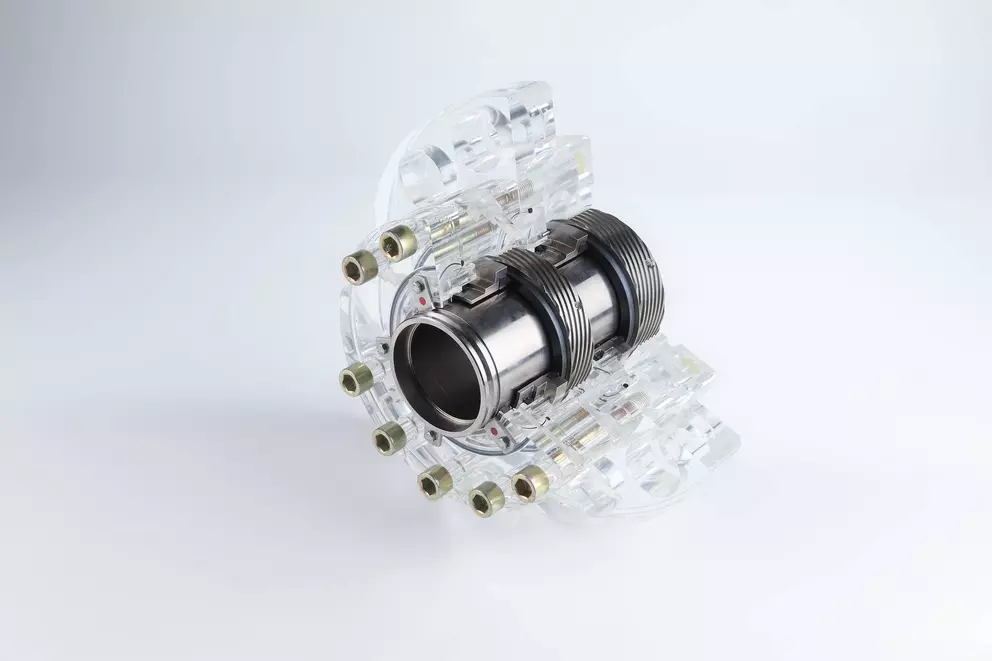 Ein mechanisches Bauteil, bestehend aus einem zentralen Metallzylinder mit einem gewellten Abschnitt, umgeben von einem segmentierten, transparenten Acrylgehäuse, das durch mehrere kleine Schrauben zusammengehalten wird und auf einer weißen Oberfläche ruh