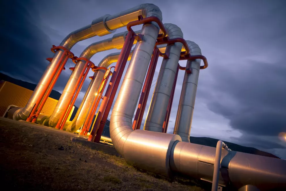 Große silberne Industrierohre mit roten Stützen wölben sich vor einem dramatischen dunkelblauen Abendhimmel nach oben.