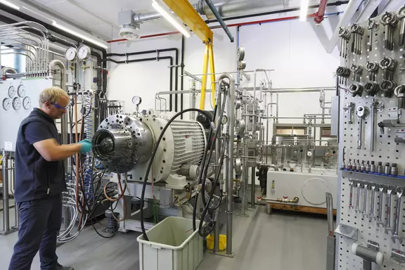 Techniker arbeitet in einer industriellen Werkstatt und überprüft oder montiert ein großes Pumpen- bzw. Dichtungssystem, umgeben von Werkzeugen und Rohrleitungen.