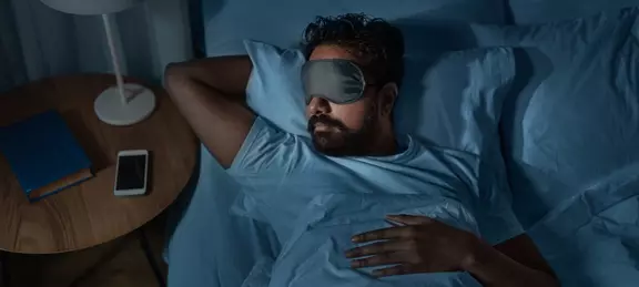 A man with a beard is sleeping in bed wearing a gray eye mask, with his head on a pillow and his arm behind his head, under dark blue sheets. A smartphone and a book are visible on the nearby nightstand under a soft lamp.
