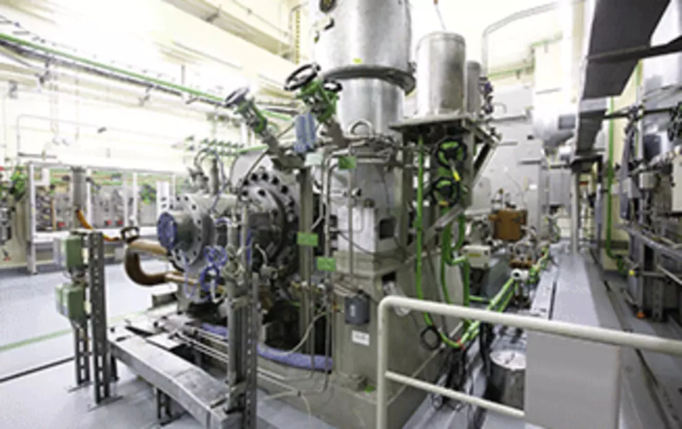 A bright, wide-angle photo of a large, complex industrial turbo-compressor or turbine machine, with numerous pipes and gauges, situated in a clean, brightly lit industrial facility.