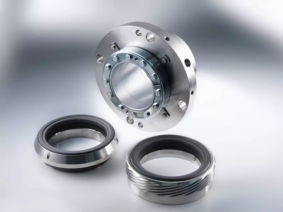 A studio photograph showing a disassembled industrial mechanical seal, featuring a large, polished metal flange and two separate, smaller circular sealing rings with black rubber elements.