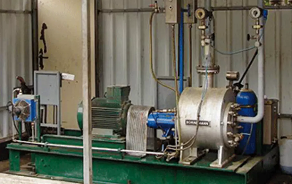 An industrial pump or compressor skid, consisting of a green electric motor connected to a silver processing unit with pipes and gauges, mounted on a dark green base against a corrugated metal wall.