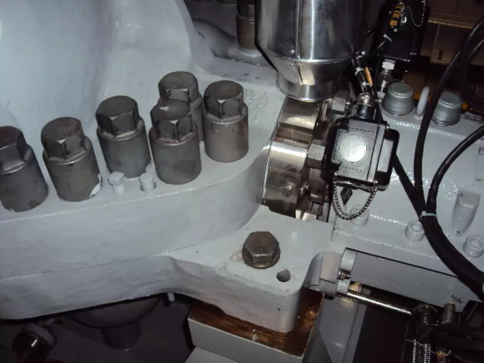 A close-up of a large industrial machine's casing, showing a cluster of large hexagonal steel bolts and a smaller sensor or monitoring device attached near the flange connection.