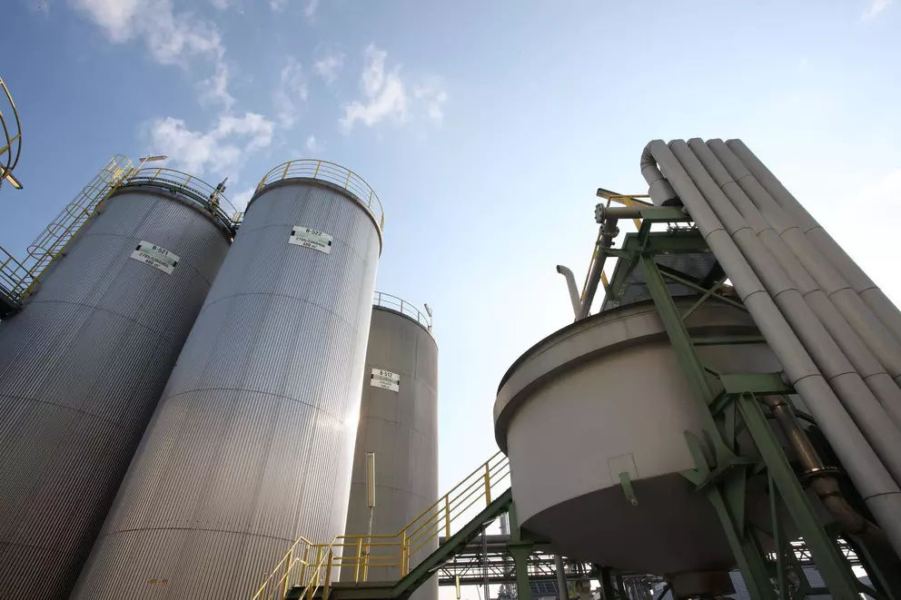 A striking low-angle view of three tall, corrugated metal storage silos and a large industrial tank with multiple vertical pipes, set against a bright, slightly cloudy blue sky. Yellow railings and ladders are visible on the structures.