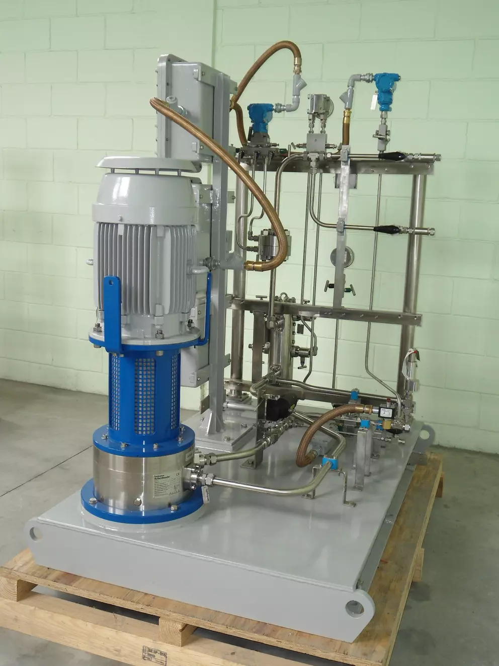 A close-up of industrial machinery, likely a lube oil or hydraulic power unit, featuring a large grey and blue electric motor and pump assembly connected to a stainless steel piping and valve rack, all mounted on a grey skid base on a wooden pallet.
