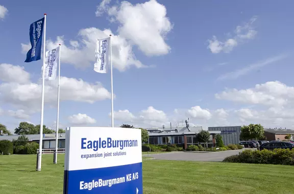 EagleBurgmann KE A/S-anlæg i Danmark med firmaskilt, flag og kontorbygningens ydre, der fremhæver stedets tilstedeværelse og virksomhedens identitet.