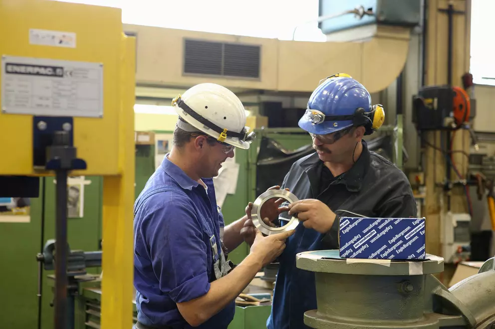 Two industrial workers in hard hats and safety gear are standing in a workshop, examining a metal ring component between them. A box labeled "EagleBurgmann" rests on a machine part in the foreground.