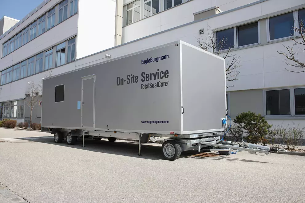 A large, silver mobile service trailer labeled "EagleBurgmann On-Site Service TotalSealCare™" is parked on asphalt outside a modern, white industrial or office building.