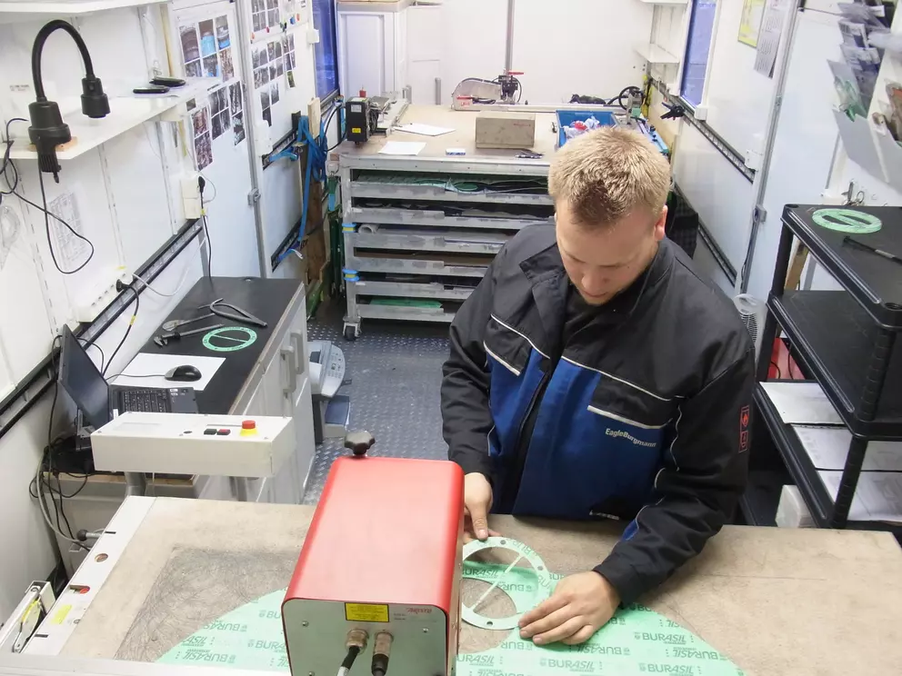 A worker in a blue and black uniform is examining a green circular gasket being cut by an industrial machine with a red housing, inside the confined space of a mobile service workshop.