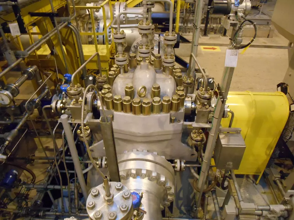A high-angle view of a large, complex industrial machine, likely a centrifugal compressor or pump, featuring a thick gray casing with numerous brass-colored bolts and fittings, surrounded by pipes and yellow safety structures.