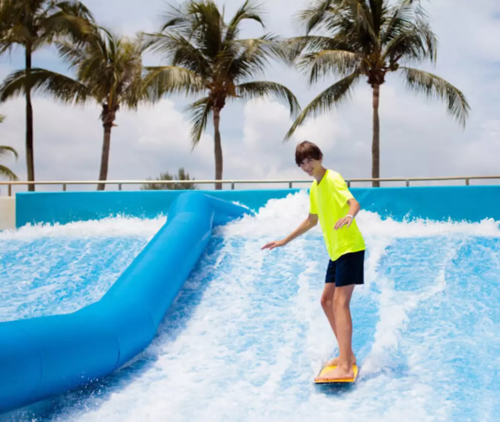 Jugendlicher beim Surfen auf einer künstlichen Welle in einem tropischen Wasserpark mit Palmen im Hintergrund.