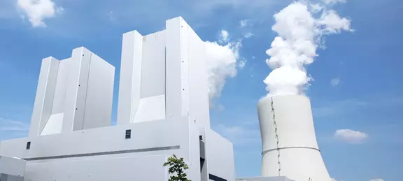 A striking, low-angle photo of two large, modern, white rectangular industrial buildings next to a massive cooling tower emitting white steam, all set against a blue sky with scattered clouds.