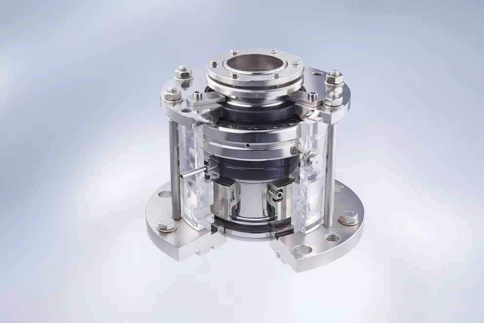 A studio shot of a high-precision, metallic mechanical seal assembly, mounted between two large flanges, with the internal components visible through a partially cutaway clear acrylic housing.