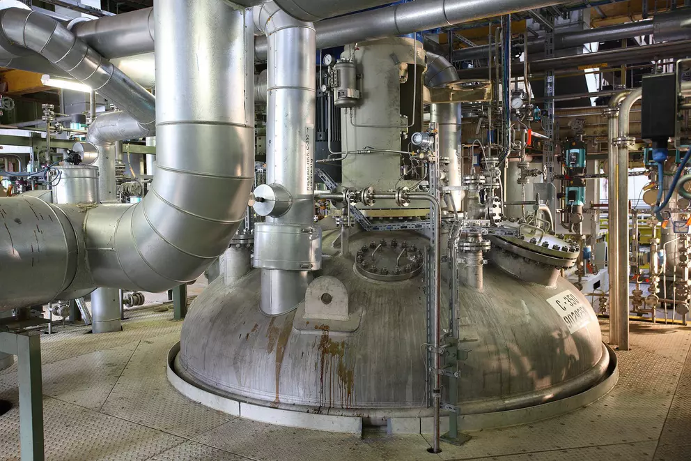 A large, domed industrial reactor or mixing vessel in a factory, surrounded by complex silver piping and instrumentation, with visible signs of wear and fluid staining.