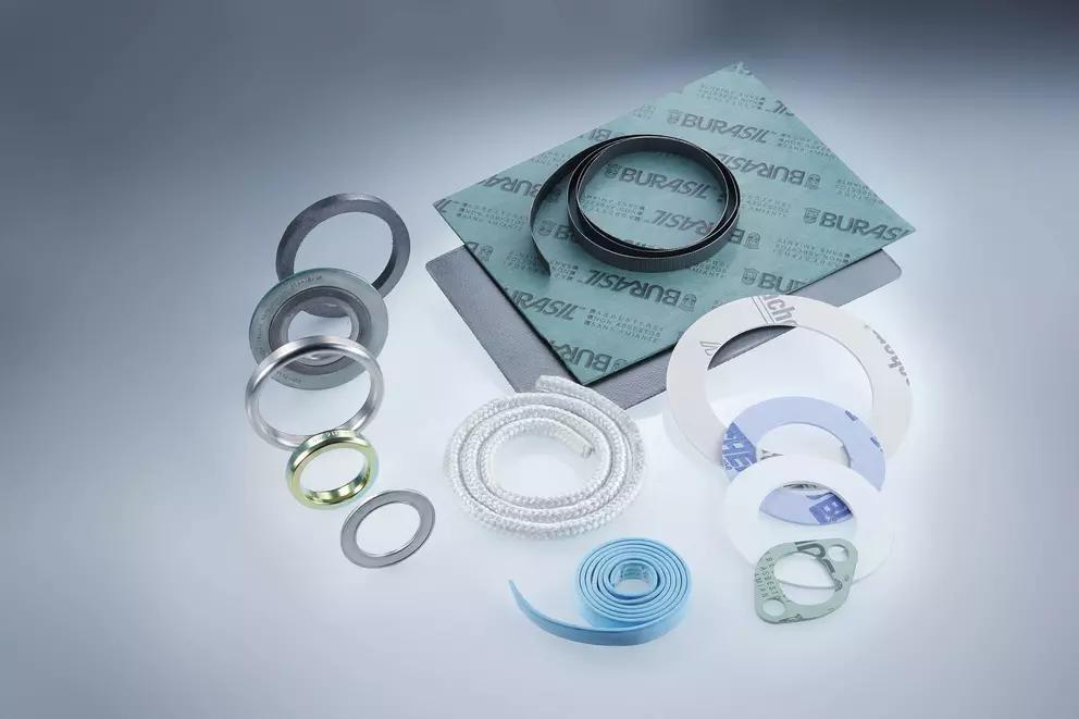 Eine Sammlung von Dichtungs- und Dichtungsmaterialien, darunter gewalzte weiße Geflechtpackungen, Metallringe, flexible blaue Streifen und Platten aus „BURASIL”-Faserdichtungsmaterial.
