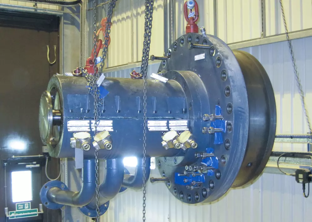 Ein großes, dunkles industrielles Maschinenbauteil, möglicherweise ein Teil einer Hochleistungspumpe oder -turbine mit mehreren Schraubverbindungen und angeschlossenen Rohrleitungen, hängt in einer Fabrikumgebung an dicken Metallketten.