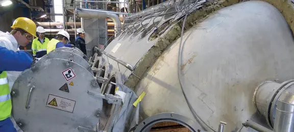 Trabajadores con cascos y ropa de seguridad inspeccionan un gran equipo cilíndrico industrial en una planta, rodeado de tuberías y componentes metálicos.