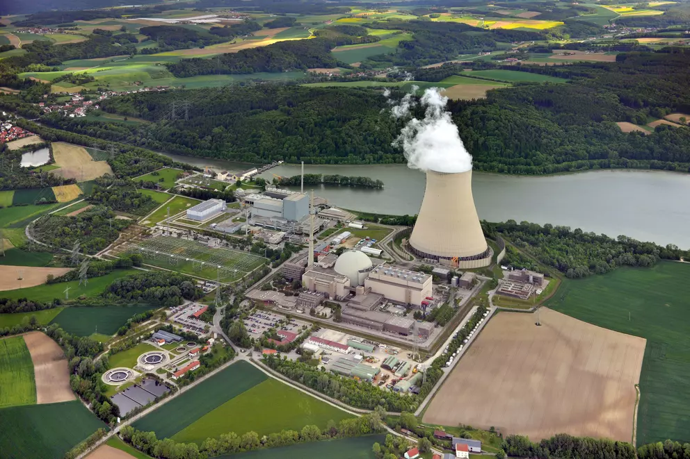 An aerial view of a nuclear power plant complex located by a large body of water, surrounded by mixed agricultural fields and forests. A single cooling tower is visible, emitting a large plume of white steam.
