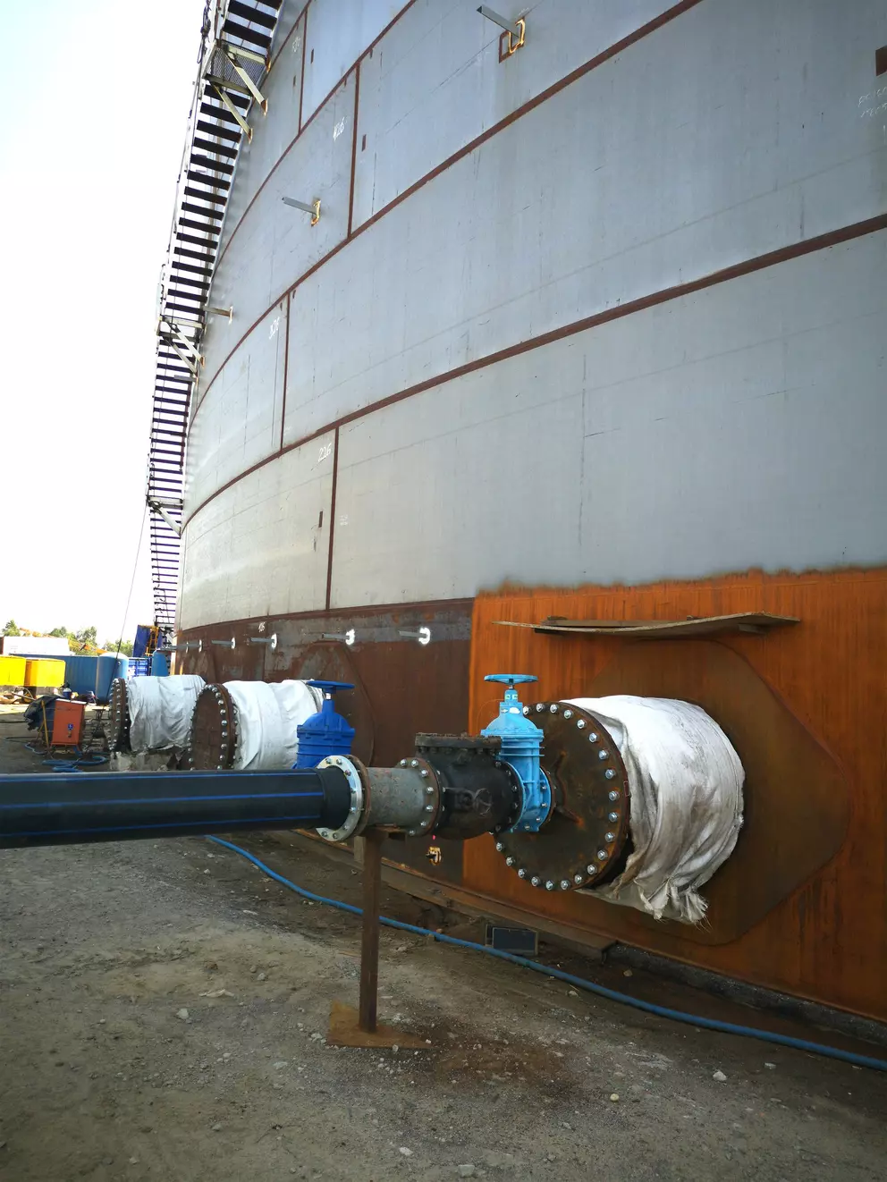 A ground-level view of a large, cylindrical industrial storage tank under construction. Several large pipes with blue valves and flanges are connected to the base of the tank.