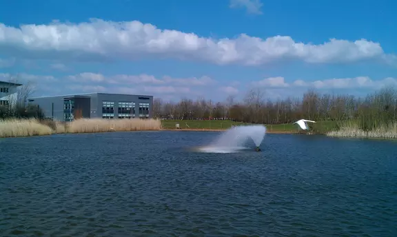 EagleBurgmann office building beside a landscaped lake with fountain under a blue sky with scattered clouds.