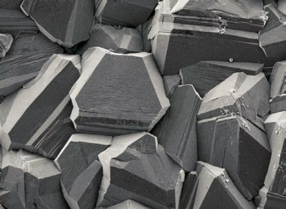 Nahaufnahme von Kristallstrukturen unter dem Mikroskop, die scharfkantige, geometrische Formen mit glatten Oberflächen und eckigen Facetten in Graustufen zeigen.