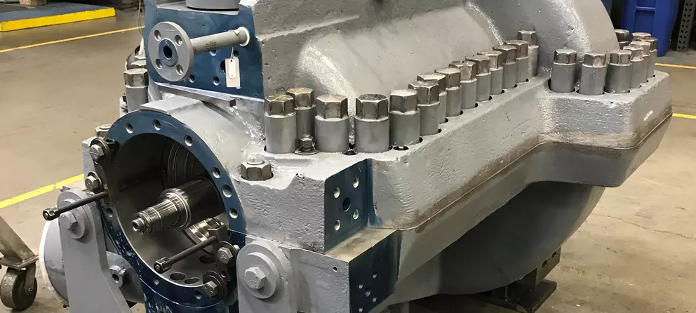 A large industrial centrifugal pump partially disassembled in a workshop setting, showing the internal shaft and components. The pump housing is gray with a series of large bolts on top, and the surrounding area appears clean and well-lit.