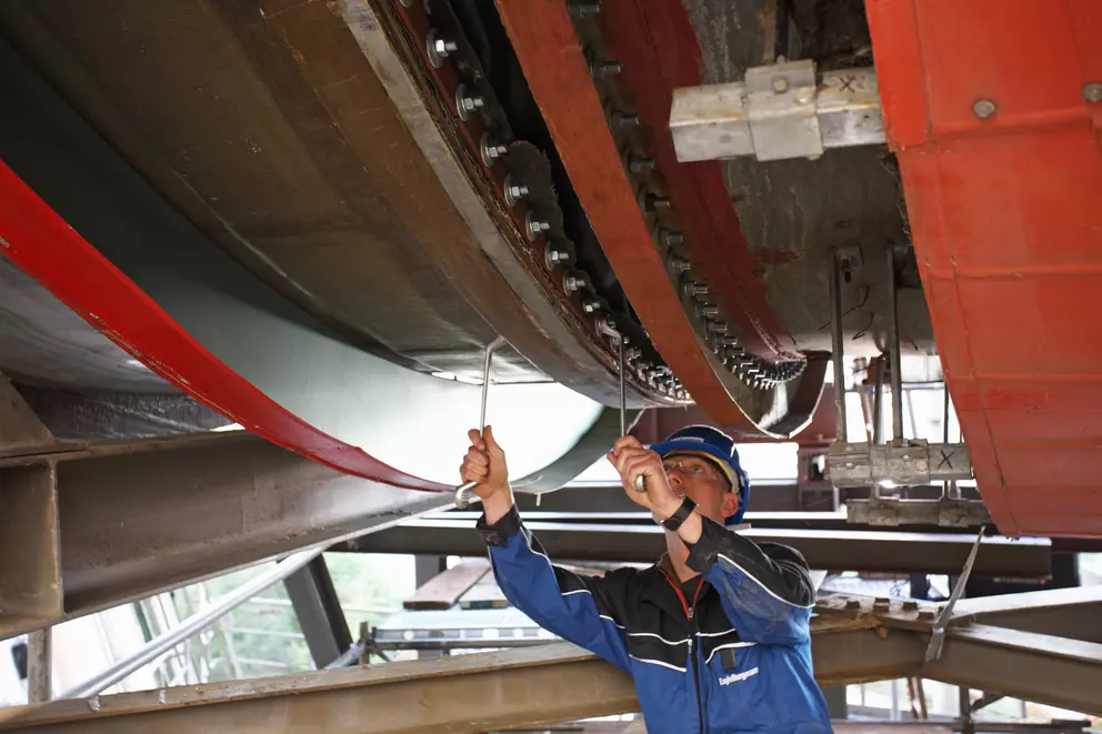 A worker in a hard hat and blue jacket is reaching up to tighten a long row of bolts on a massive industrial structure, which features red and brown bolted beams overhead.