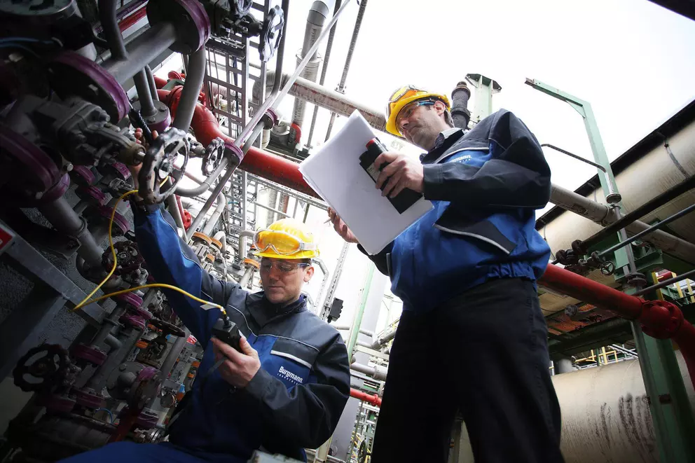 Two male industrial workers in hard hats and safety glasses are performing maintenance on complex machinery and piping in a factory. One worker is checking instruments on a valve assembly, while the other stands above, holding a clipboard and looking at a
