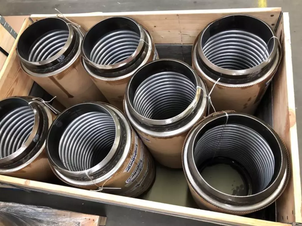 A wooden crate holds several large, cylindrical objects. Each object consists of a tightly coiled metal spring or tube inside a cardboard and metal casing.