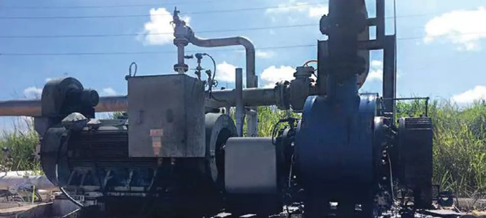 A photo of a large, dark industrial pump and motor unit, possibly for oil or gas, stained with a dark substance, sitting outdoors amongst tall grass and under power lines.