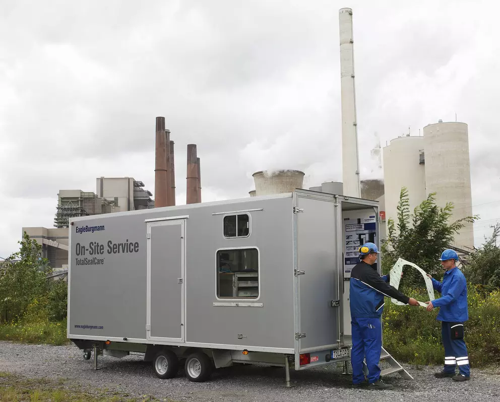 A silver mobile service trailer labeled "EagleBurgmann On-Site Service" is parked in a gravel lot outside an industrial facility with tall chimneys and a cooling tower. Two workers in blue coveralls are standing at the open rear of the trailer, examining