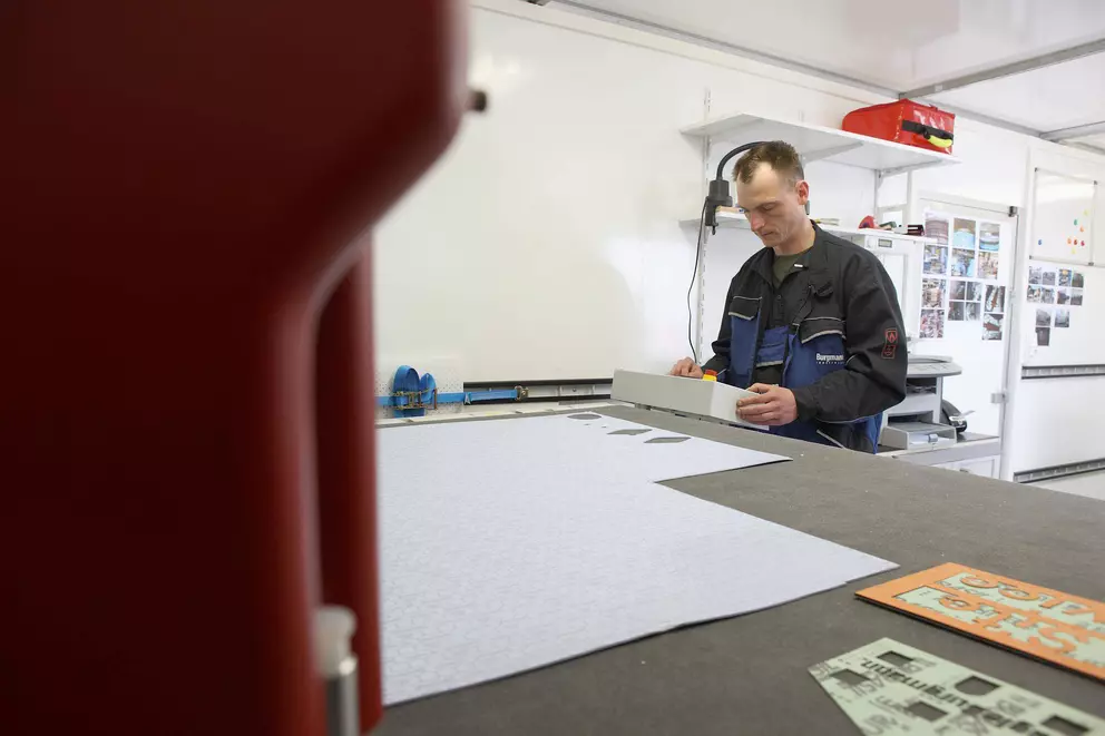 A male worker in blue and black overalls is standing at a large workbench in a brightly lit industrial workshop, examining a long, white rectangular component placed on a sheet of gasket material.