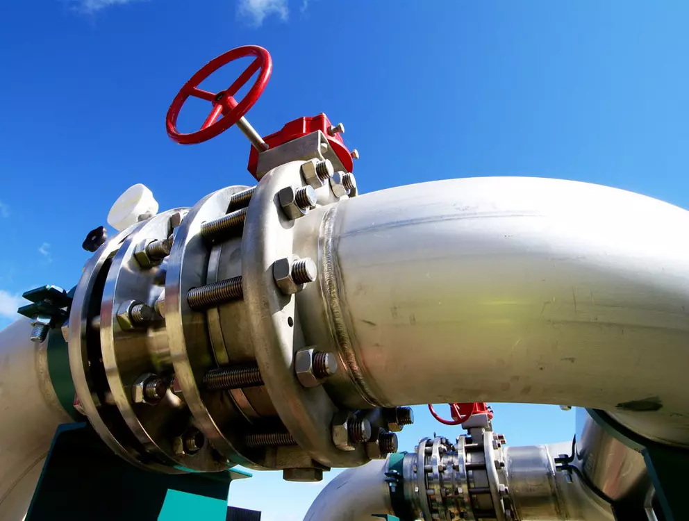 A low-angle shot of a large industrial metal pipeline, featuring a heavily bolted flange connection and a valve with a bright red handwheel, set against a clear blue sky.