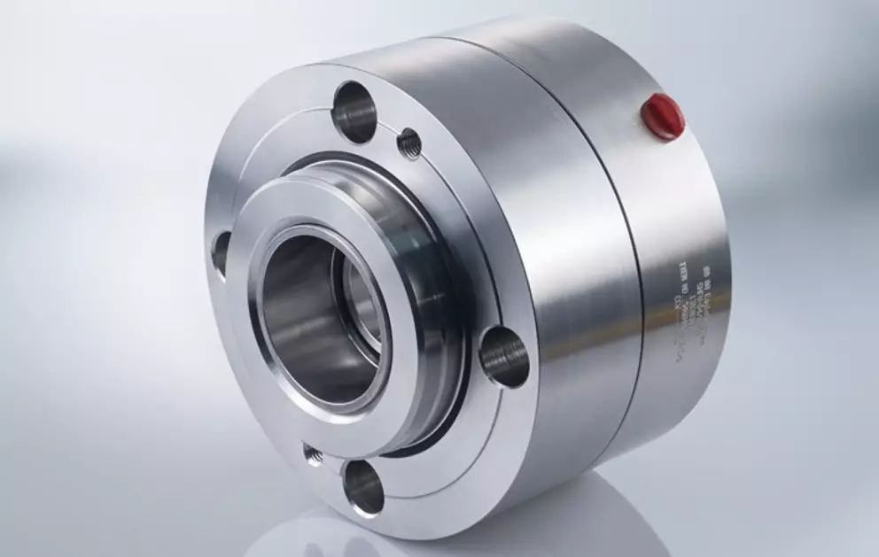 A professional studio photograph of a shiny, stainless steel industrial mechanical seal or coupling component, featuring a cylindrical body with bolt holes and a central opening, with a small red cap visible on the side.