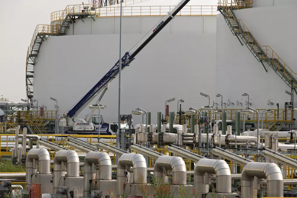 A large white storage tank at an industrial facility is visible behind a dense foreground of shiny, curved pipelines and a crane.