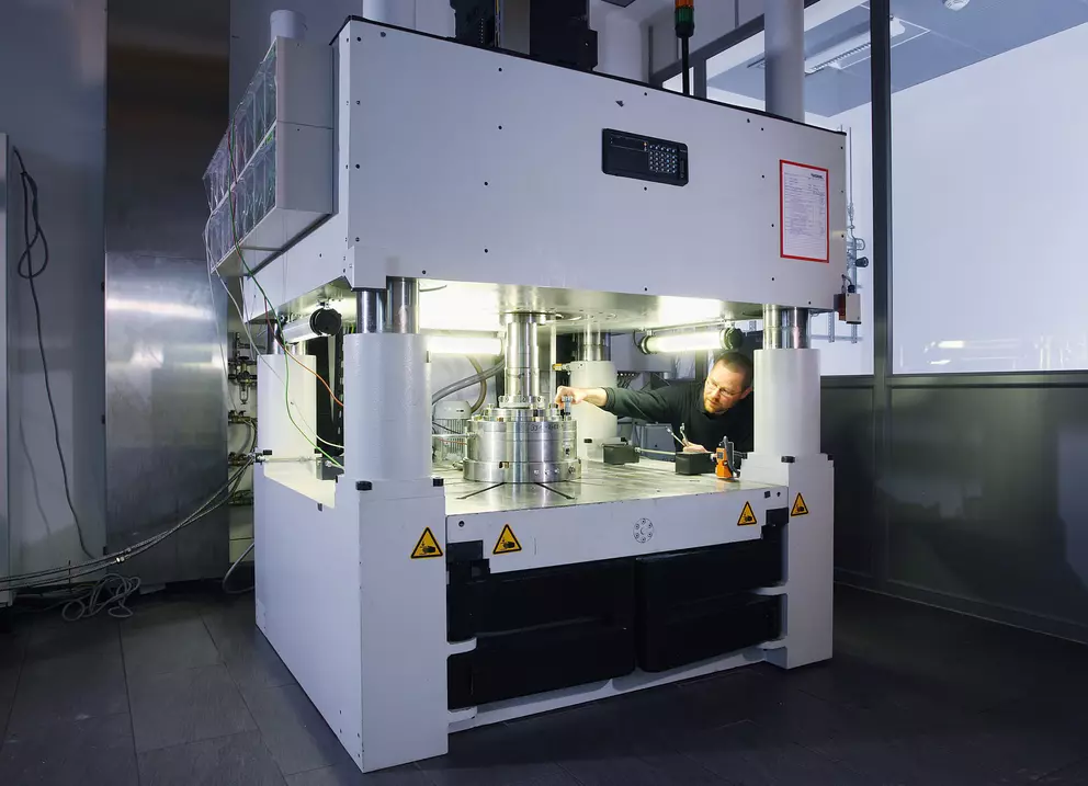 Technician working on a large, white industrial testing or pressing machine with bright internal lighting in a laboratory setting.