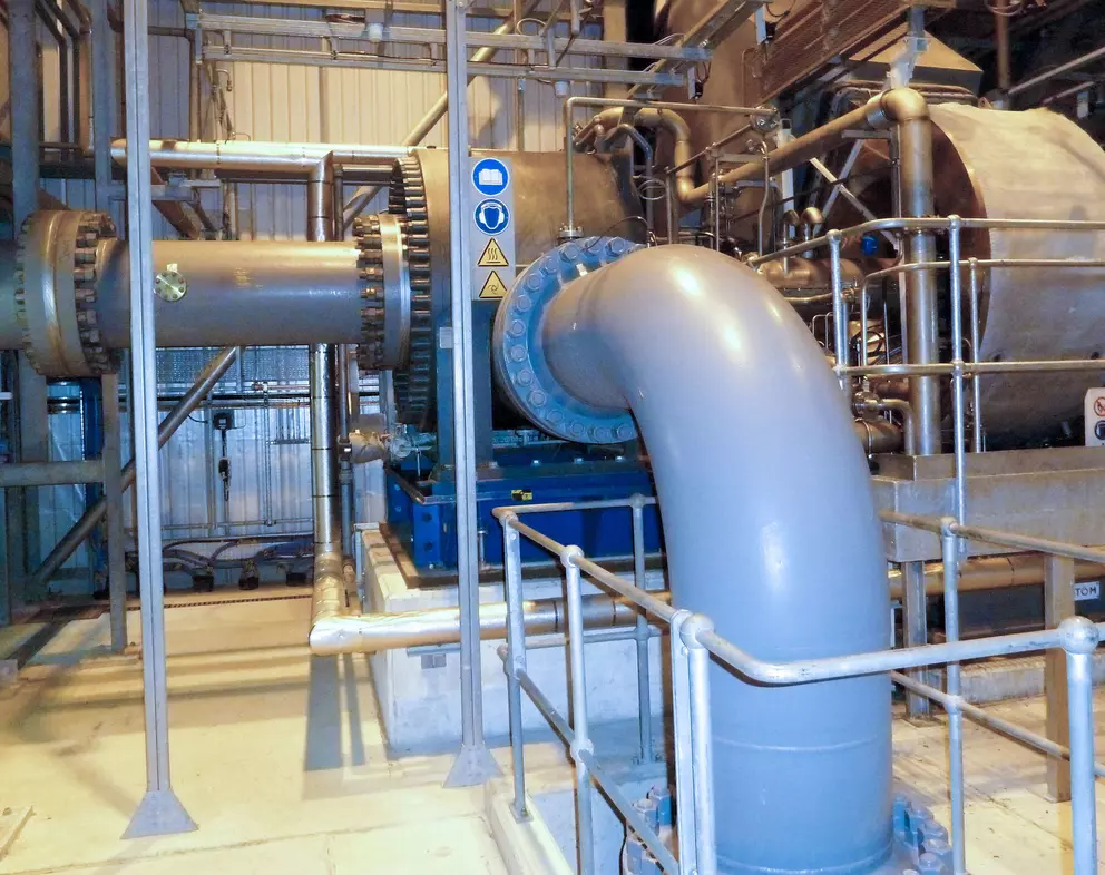 Ein Blick in einen Industrieinnenraum, der eine große horizontale Maschine zeigt, möglicherweise ein Getriebe oder eine Turbine, die mit einem massiven, grauen, gebogenen Rohr verbunden ist und von Sicherheitsgeländern und Rohrleitungen umgeben ist.