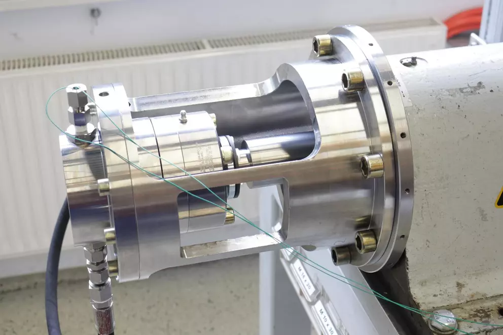 A polished stainless steel industrial component, likely a mechanical seal or pump component, with an exposed cross-section showing its internal construction with bearings and a central shaft on an industrial test rig.