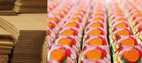 Montones de cartón apilado junto a filas de envases de cartón para bebidas con tapones naranjas, mostrando materiales de embalaje y producción industrial.