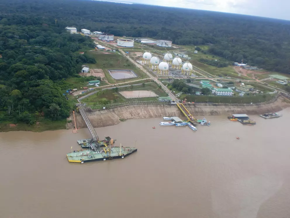 Aerial view of an oil and gas facility by a river, with storage tanks, buildings, and boats, surrounded by dense rainforest.