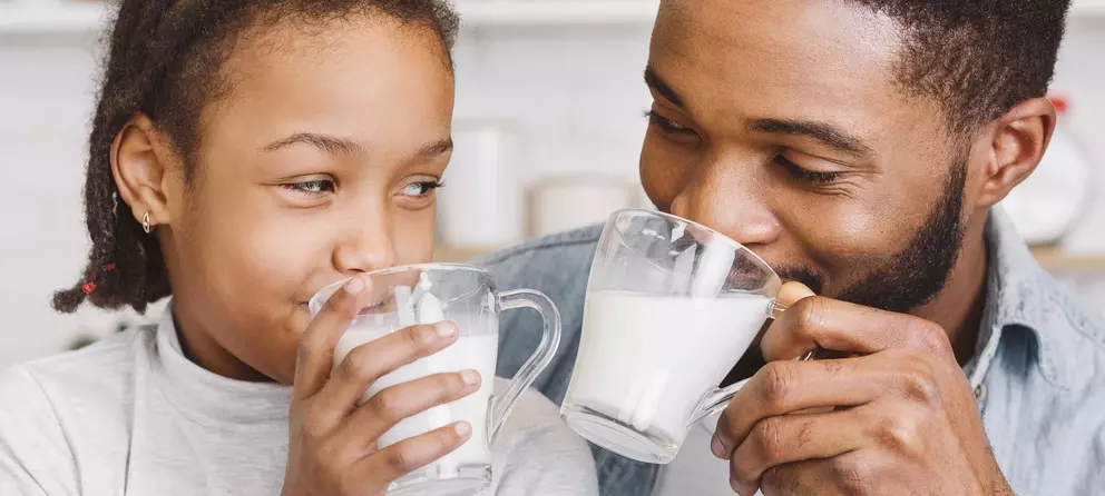 A father and daughter are smiling at each other while holding cups of milk. They appear to be enjoying a moment together, sharing a joyful and intimate interaction in a cozy indoor setting.