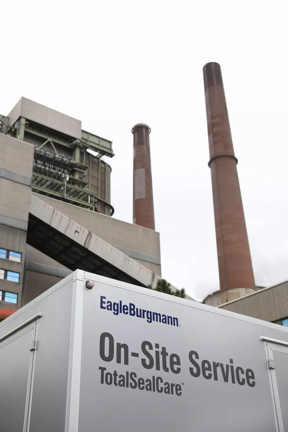 A low-angle shot showing the top corner of a silver service trailer labeled "EagleBurgmann On-Site Service TotalSealCare™" in the foreground, with the large, imposing structure and two tall brown chimneys of an industrial power plant rising in the backgr