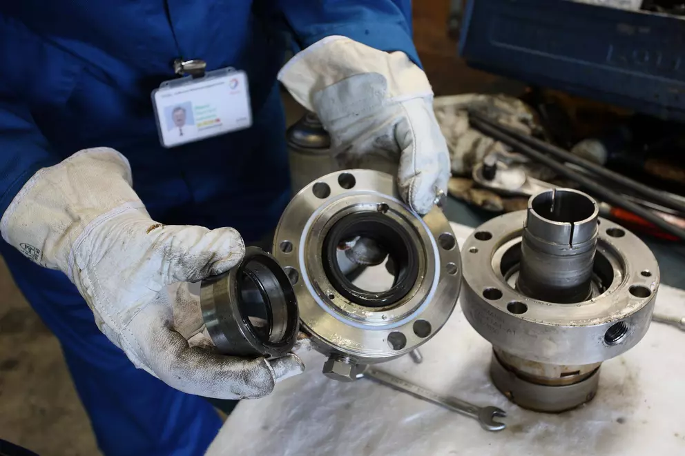 A close-up of a worker in white gloves holding two disassembled parts of a mechanical seal: a small black seal ring and a larger metal flange component, with another seal part resting on a workbench in the background.