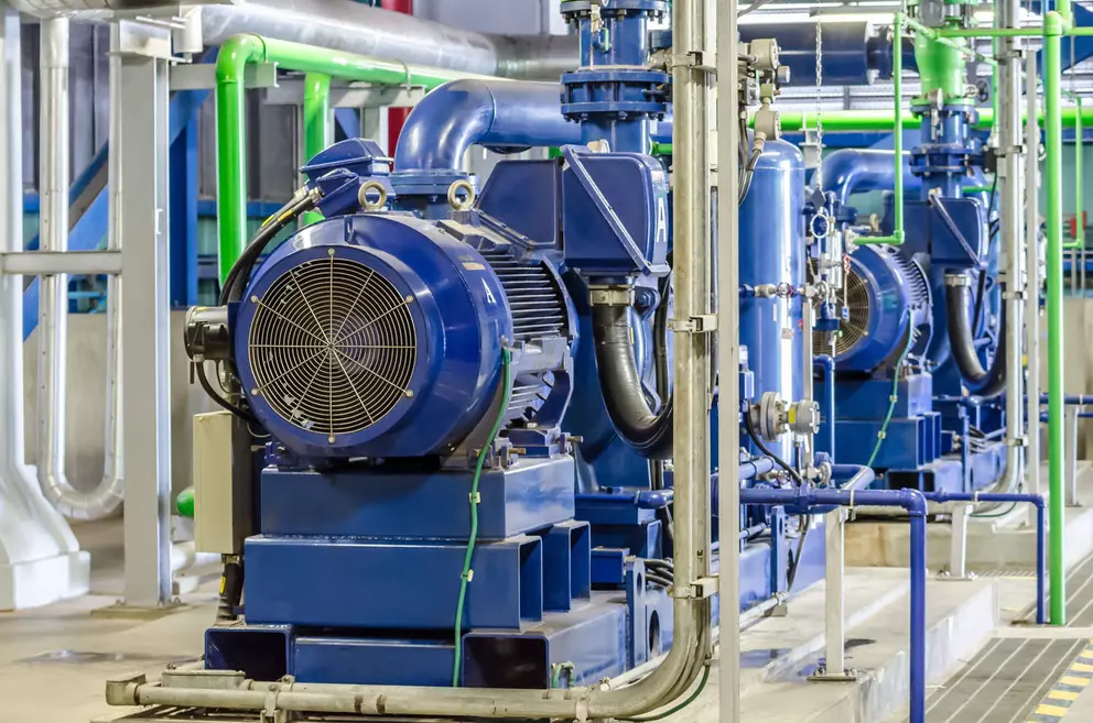A row of large, bright blue industrial electric motors and pumps connected to a network of pipes, including some painted green, within a clean industrial facility.