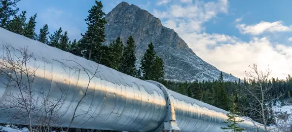 A large, silver industrial pipeline covered in snow, stretching across a snowy landscape. The pipeline runs diagonally from the bottom left to the center right of the image, with a snow-covered evergreen forest and a large, rocky mountain peak in the bac