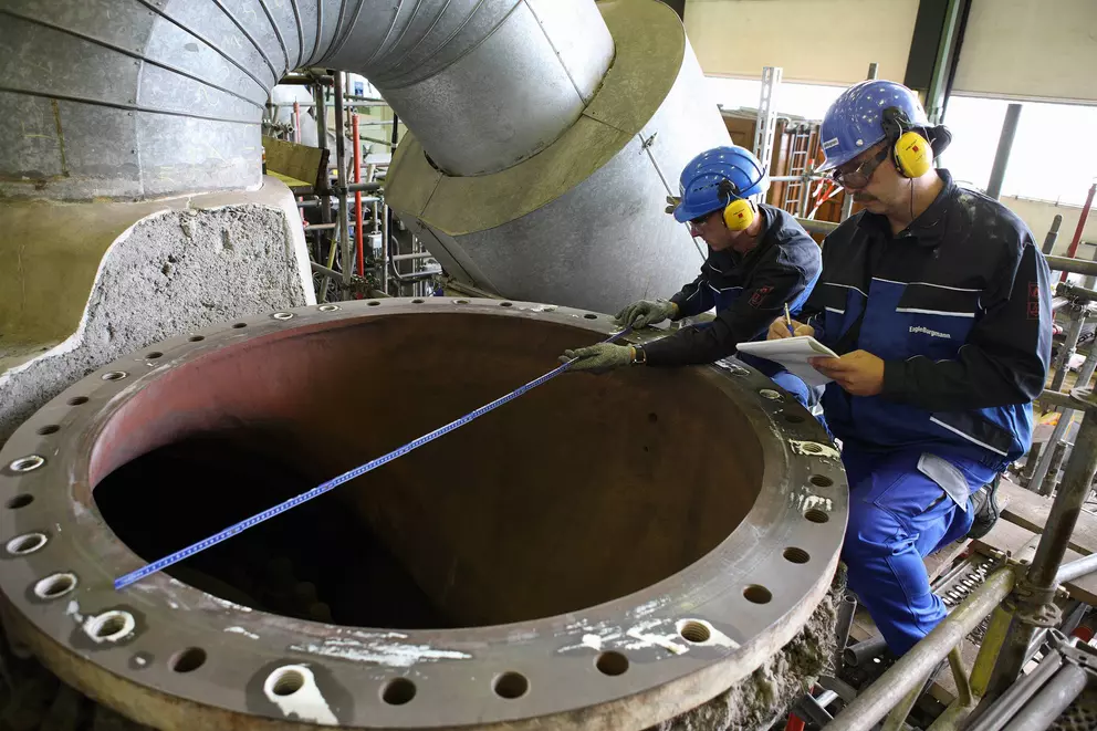Two industrial technicians in hard hats and safety gear are taking measurements of a large, open flange on a section of industrial piping using a blue measuring tape, with one worker recording notes.